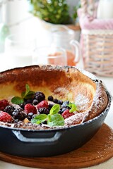 Fresh homemade dutch baby pancake with powdered sugar, fresh berries and mint for breakfast with a glass of milk and apple juice on a light background. Delicious sweet breakfast in a sunny interior. 