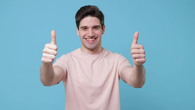 Smiling cheerful young man 20s years old wearing casual beige t-shirt posing isolated on pastel blue colour background in studio. People emotions lifestyle concept. Looking camera showing thumbs up
