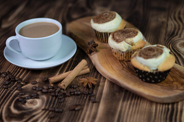 A white cup of coffee with homemade cinnamon cupcakes on a wooden board on a textured brown table composition closeup 