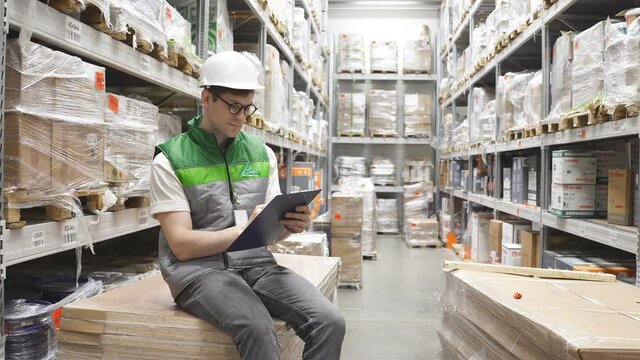 Warehouse inventory management. Warehouse worker is writing, keeping count, caucasian guy in helmet and uniform is concentrated on work in warehouse.