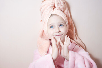 a little girl in a pink dressing gown with a towel wrapped around her head smears her mother's cream on her face. pink background