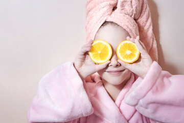 a little girl in a pink robe and a bath towel on her head covers her eyes with oranges on a pink background