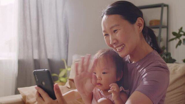 Happy Asian woman sitting on sofa with cute baby daughter, smiling and waving at smartphone while video calling from home