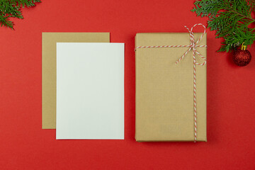 Christmas letter concept. Blank paper card, craft paper envelope, vintage gift box, rope, thuja branches on red background.Holiday Flat lay. Top view. Copy space. Mockup.