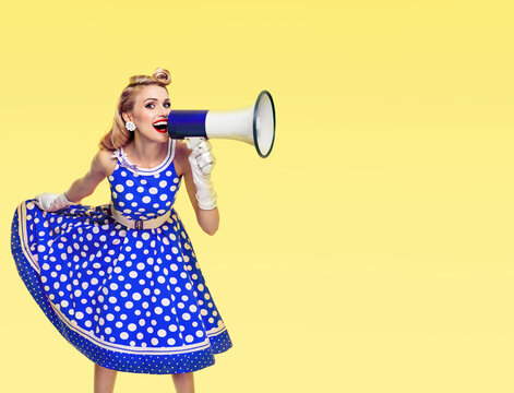 Happy excited woman holding megaphone, dressed in pin up style blue dress in polka dot and white gloves, on yellow background. Beautiful blond girl in retro fashion vintage studio picture.