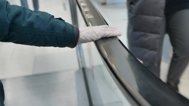 Hand in white protective medical glove holds and slides on long black escalator handrail in shopping mall closeup slow motion