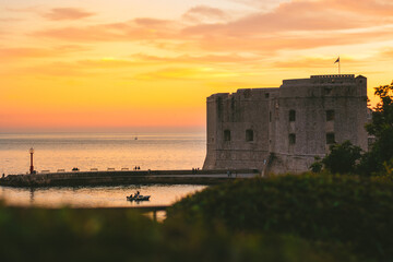 Golden sunset in the town of Dubrovnik, castle standing on the shore of a small harbour, sailboat leaving the harbour and a small local fishing boat entering. Tranquil simple serene setting
