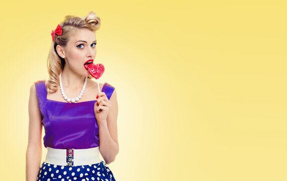 Woman eating heart shape lollipop dressed in pinup style dress, with copyspace area for slogan or advertising text message, on yellow background. Pinup girl in retro fashion and vintage studio concept