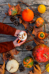 Coffee latte with whipped cream and cinnamon on top held in womans hands on a wooden background of pumpkins, autumn leaves. Fall and Thanksgiving concept. Top View