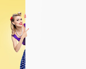 Very happy smiling woman in pin-up style dress, showing blank signboard with copy space, over yellow background. Pinup blond girl posing in retro and vintage studio concept.
