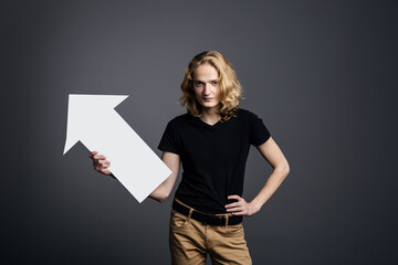 A handsome young guy with long blonde hair and a smile face holds a white arrow pointing to the right on a gray background