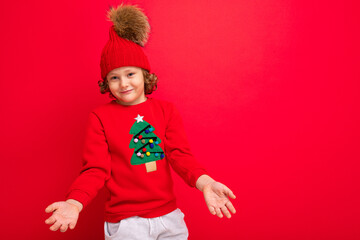 cool teenager in a red Christmas sweater fooling around against the background of a red wall, a warm hat and a sweater with a Christmas tree