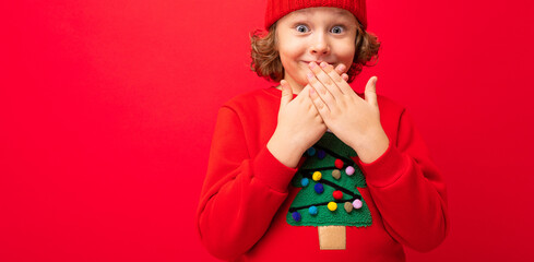 cool teenager in a red Christmas sweater fooling around against the background of a red wall, a warm hat and a sweater with a Christmas tree