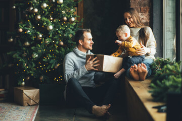 Fototapeta na wymiar A happy family of three sits at home near festive Christmas tree. Young parents and little child smile. New Year mood