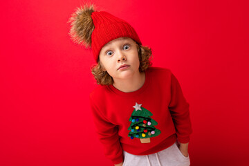 cute blond boy in warm hat and christmas sweater on red background with smile on his face