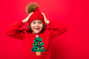 cute blond boy in warm hat and christmas sweater on red background with smile on his face