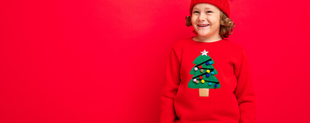 cute blond boy in warm hat and christmas sweater on red background with smile on his face