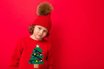 cool blond kid in warm hat and sweater with christmas tree on red background fooling around, christmas concept