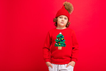 cute blond boy in warm hat and christmas sweater on red background with smile on his face
