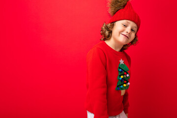 cute blond boy in warm hat and christmas sweater on red background with smile on his face