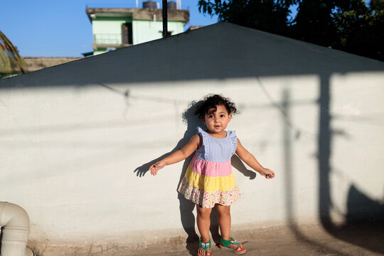 Cute little baby girl standing against a wall wearing colorful frock in direct afternoon sunlight