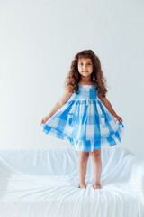 Beautiful little curly girl in a blue dress on the white couch in the room