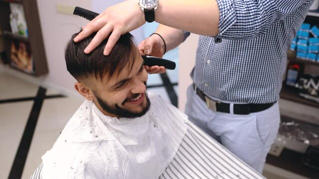 Young man in barber shop sits in an armchair. Barber cuts his hair with a trimer