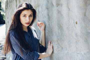 Portrait of young stylish girl in front of white concrete wall in sunny day. Portrait of beautiful girl. Pretty girl with long dark hair and red lips. 