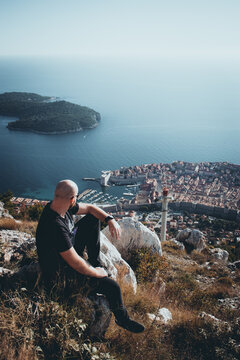 Vertical moody shot of a bald man sitting on the Srd mountain above the city of Dubrovnik, observing the area on a cold autumn day