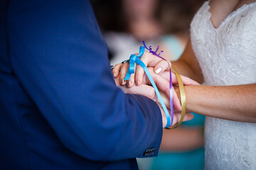 Close up picture of bride and groom's hands tied with colorful blue, purple and yellow ribbons, white lace wedding dress,  blue suit 