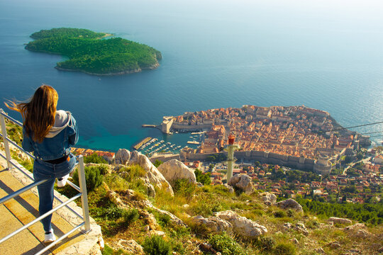 Attractive brunette traveller sitting on a railing above the city of Dubrovnik, viewpoint on Srd mountain. Looking at the ancient historic city surrounded by the blue adriatic sea