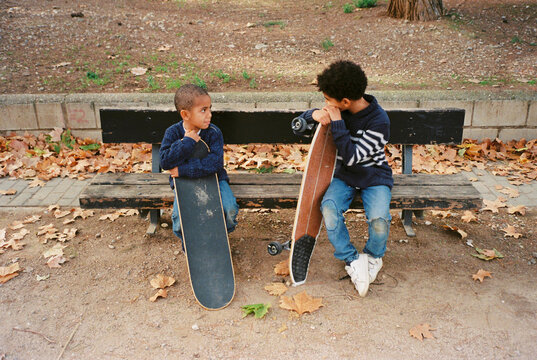 73/5000 Mixed race kids sitting on a bench in a park with their skateboards.