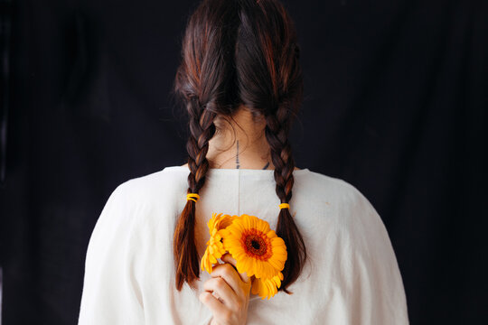 Woman from behind with braids and white shirt, she holds orange flowers behind her back