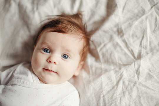 Closeup portrait of cute Caucasian red-haired newborn baby. Adorable funny child infant with blue grey eyes lying on bed looking in camera. Authentic childhood and lifestyle candid moment.