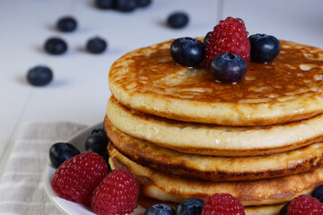 Plato de tortitas americanas con sirope de arce, arándanos y frambuesas sobre mesa de madera blanca con frutos esparcidos y desenfocados.