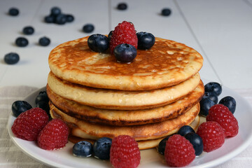 Plato de tortitas americanas con sirope de arce, arándanos y frambuesas sobre mesa de madera blanca con frutos esparcidos y desenfocados.