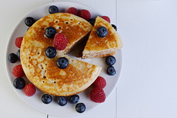 Plato de tortitas americanas cortadas con sirope de arce, arándanos y frambuesas sobre mesa de madera blanca, perspectiva cenital.