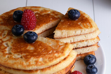 Plato de tortitas americanas cortadas con sirope de arce, arándanos y frambuesas sobre mesa de madera blanca.