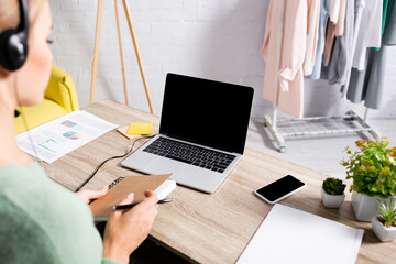 Laptop with blank screen on table near young woman in headset holding notebook on blurred foreground