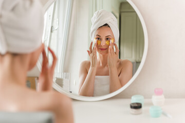 A young woman in white clothes and with a towel on her head looks in the mirror and applies gold plasters under her eyes, taking care of her skin in the morning. Woman applying gold spots on her face.