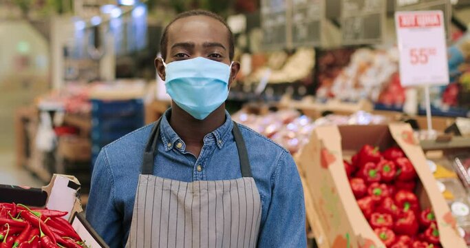 Happy young African American man seller in mask and gloves standing in supermarket with box with red hot papper and looking at camera. Male in apron working at grocery. Close up portrait concept