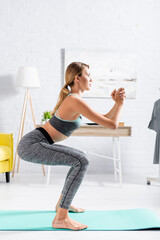 Fototapeta na wymiar Side view of young sportswoman doing squat on fitness mat at home