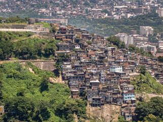 Rio de Janeiro, Brazil - December 24, 2008: Aerial view on favela built on flank of hill with brown stone cliffs. Housing on top of each other. Cityscape with green foliage in front.