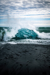 A large chunk of bright blue ice glacier piece on crystal beach in Iceland with waves crashing behind it on a black sand foreground