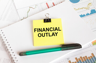 A yellow sticker with text Financial Outlay is in a Notepad with a green pen financial charts and documents