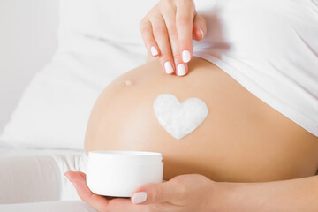 Girl sitting in bed. Young adult woman hands holding opened white cream jar. Heart shape created from cream on naked big belly. Care about perfect, soft and smooth skin in pregnancy time. Close up.
