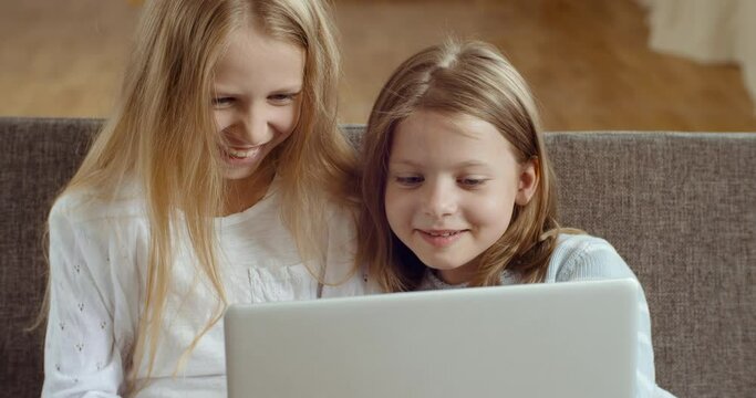 Children and technology. Two girls sisters sitting in living room together looking at laptop screen smiling laughing. Friends blonde babes watching movie cartoons, using computer for remote e-learning