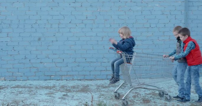 Three funny teenage boys in bright clothes ride little brother on grocery cart in backyard against blue brick wall