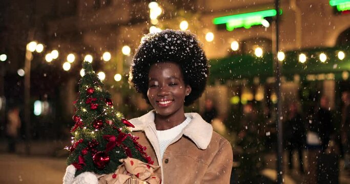Close up portrait of happy African Anerican young woman in good mood standing on street with small christmas tree looking at camera and smiling Grateful female in decorated town at night New year mood