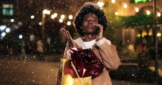 Cheerful African American woman standing with christmas gift bags and talking on smartphone while snowing outdoor. Joyful female after new years sales shopping calling on cellphone. Close up portrait